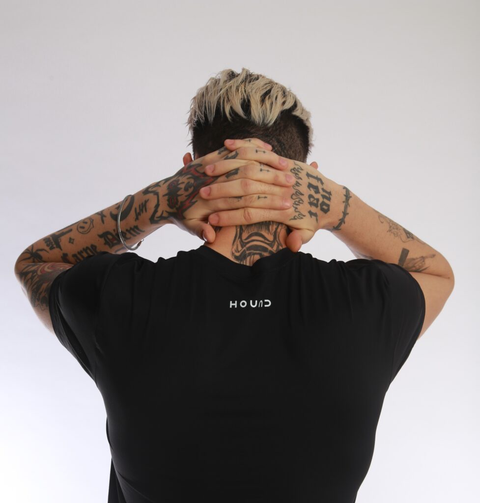 Pessoa tatuada de costas com as mãos na nuca, vestindo camiseta preta da Hound